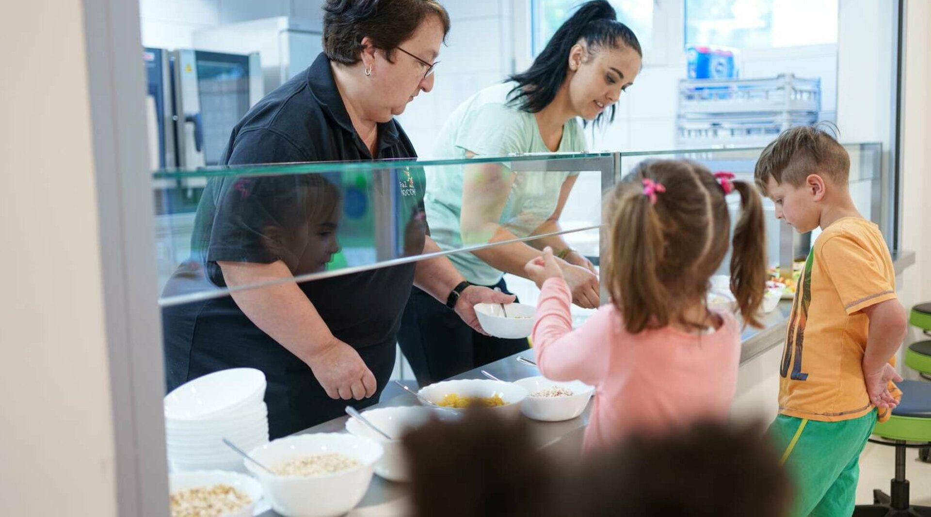 Zwei Frauen stehen hinter einer Glasvitrine und geben Essen an Kinder aus. 