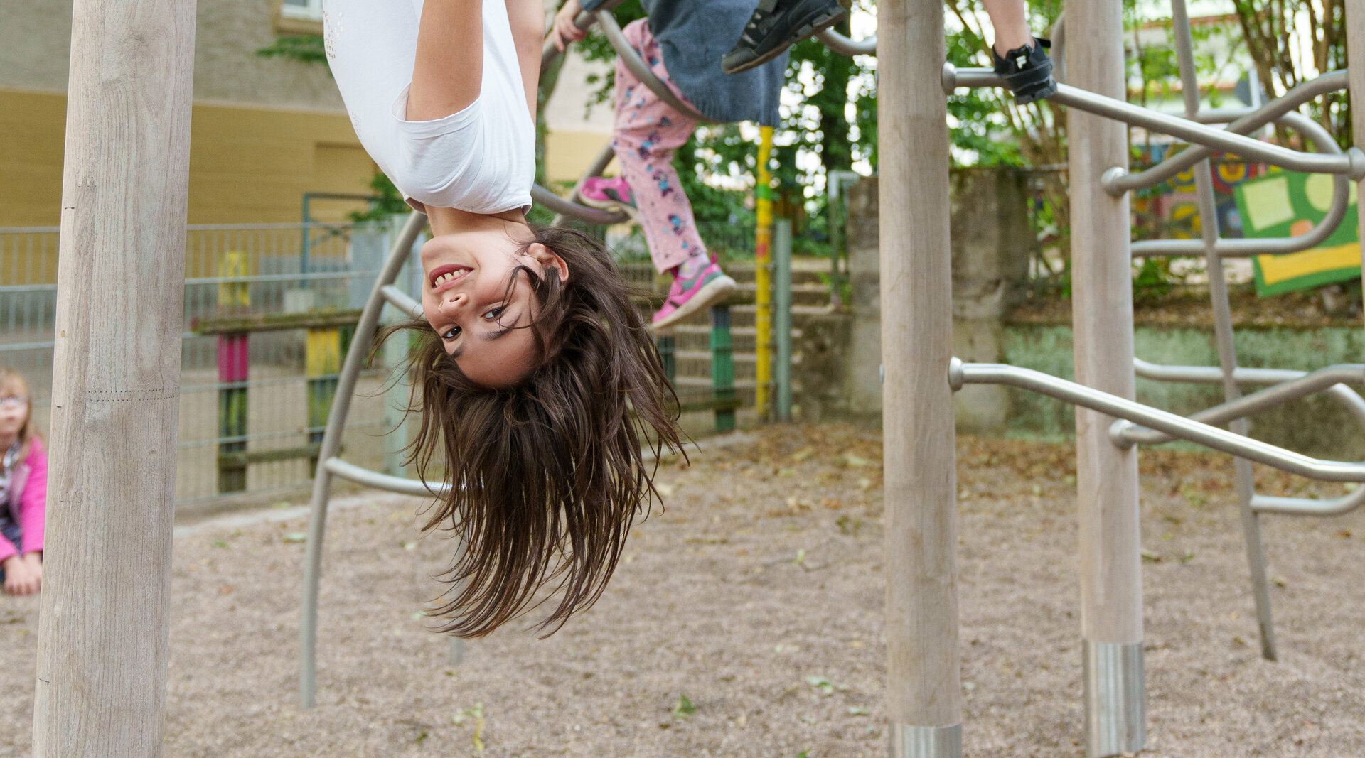 Ein Mädchen hängt kopfüber an einem Klettergerüst, das auf einem Spielplatz steht. Im Hintergrund klettert ein weiteres Kind. 